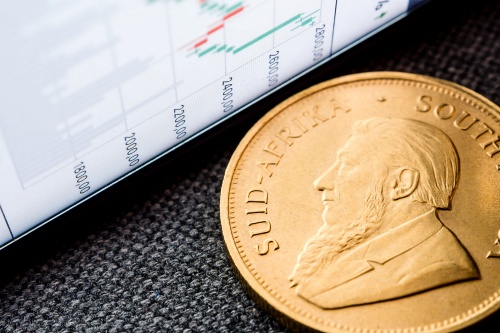 Close-up of a South African gold coin next to a tablet displaying financial charts, symbolizing investment and market tracking.