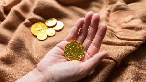 Hand holding a gold coin with additional coins resting on a soft fabric background, symbolizing wealth and precious metal investment.